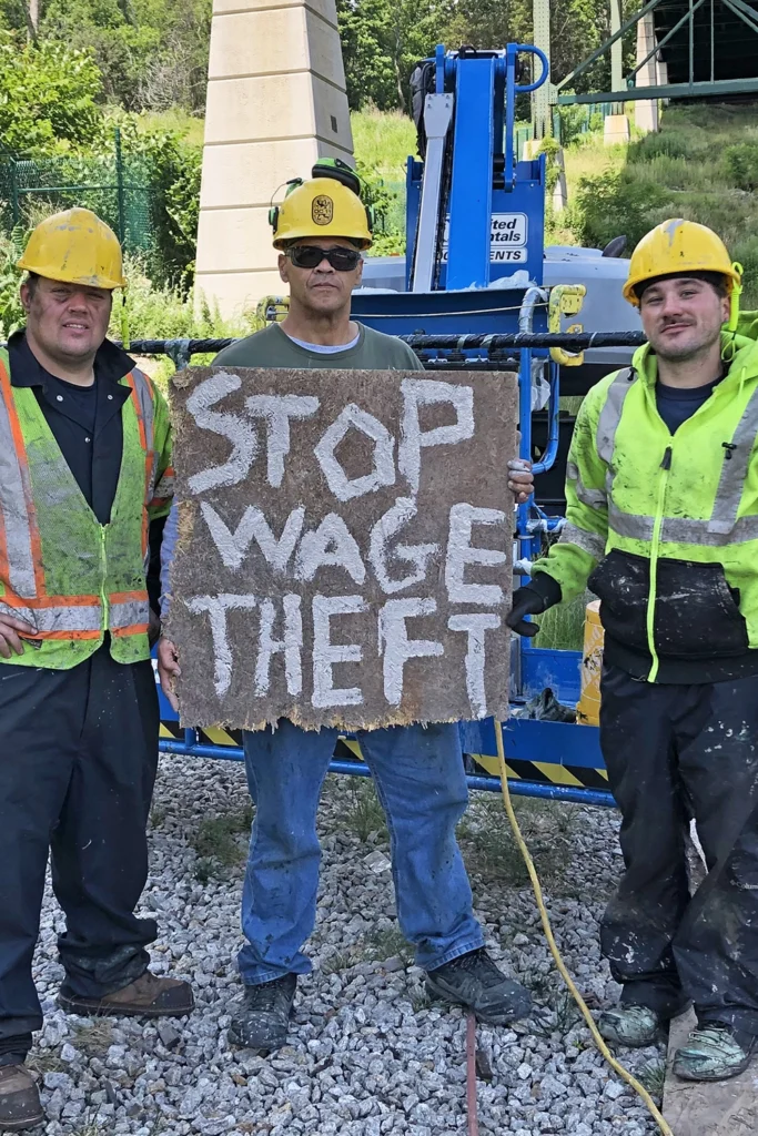 Three union construction workers wearing safety gear hold a handmade sign reading 'STOP WAGE THEFT' at a jobsite protest