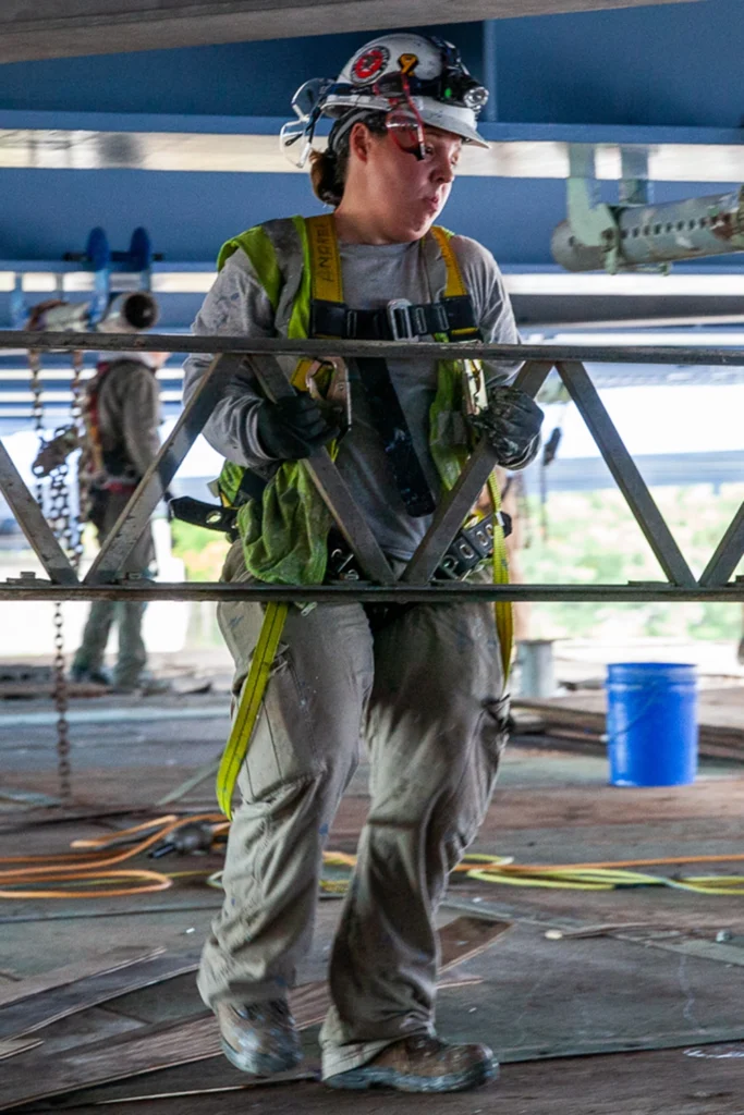 Union construction worker wearing PPE and harness while carrying metal truss on jobsite, representing IUPAT’s commitment to skilled trades and job creation